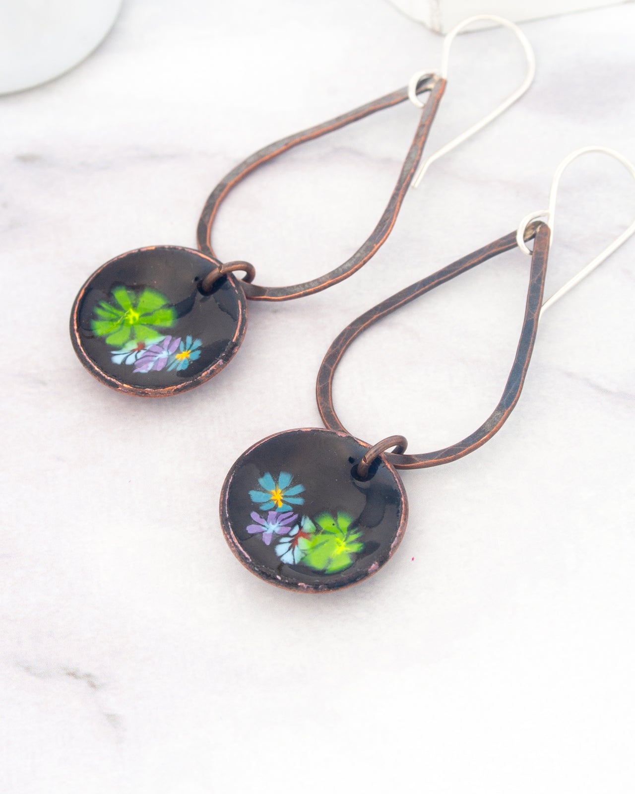 A pair of earrings with a floral design on the circular pendants, resting on a white marble surface.