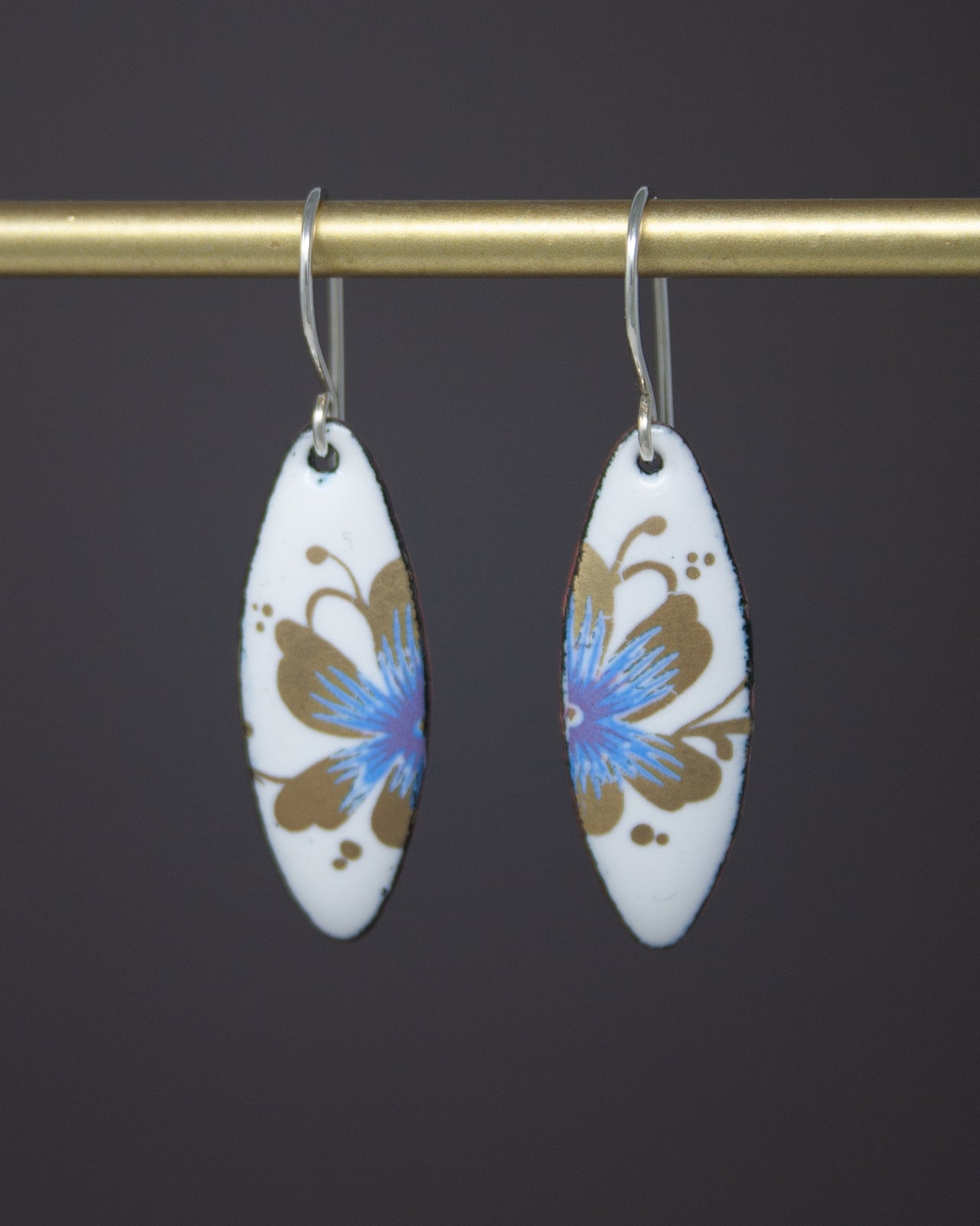 A pair of white earrings with blue and gold floral designs hanging from a gold bar.