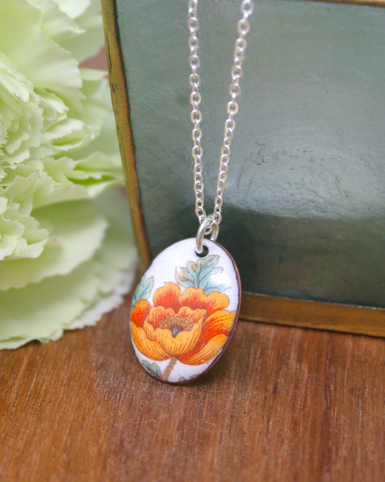 A necklace with a pendant featuring a colorful floral design is displayed on a wooden surface, with a green object in the background.