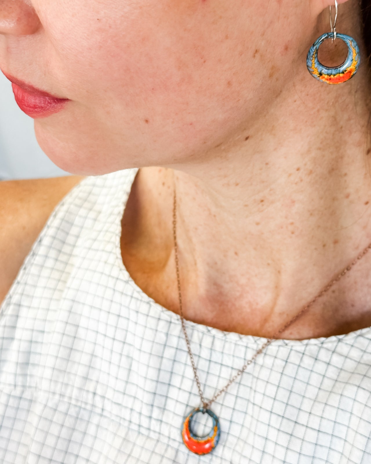 A close-up of a woman's neck and chest area, showing a necklace with a pendant and a pair of colorful earrings.