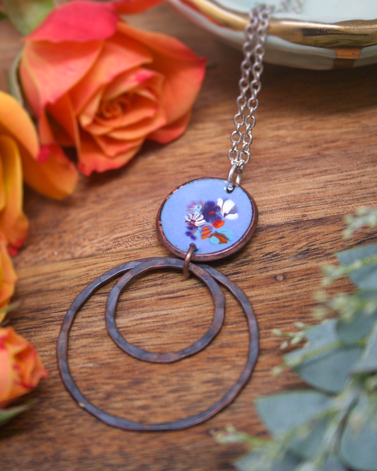 a necklace with a circular pendant, a circular ring, and a pair of circular rings, all resting on a wooden surface. There are also some orange roses and greenery in the background.