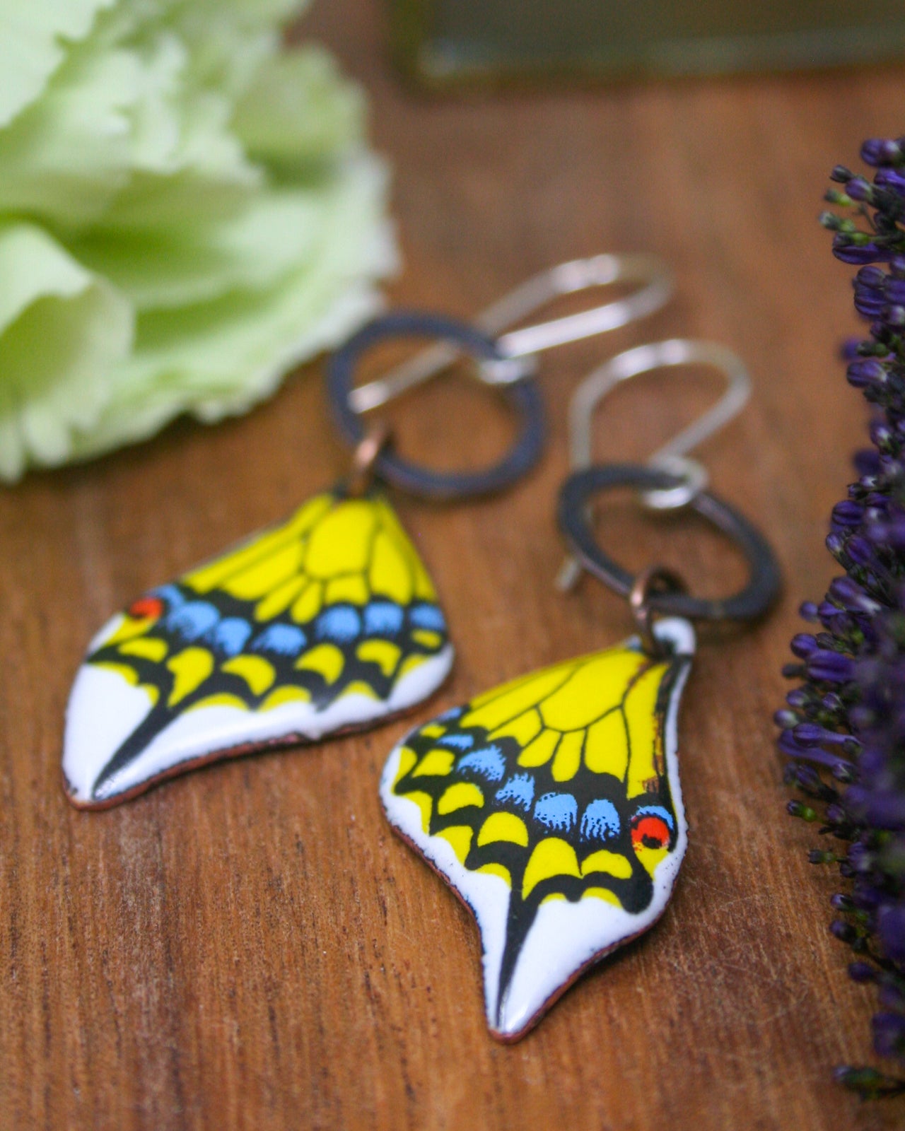 A pair of colorful butterfly-shaped earrings with intricate designs, placed on a wooden surface alongside some purple flowers.