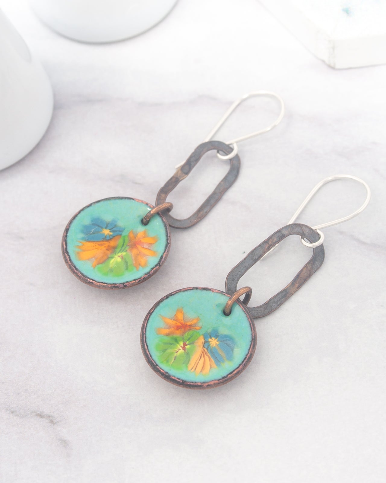 A pair of earrings with a floral design on the circular pendants, resting on a marble surface.