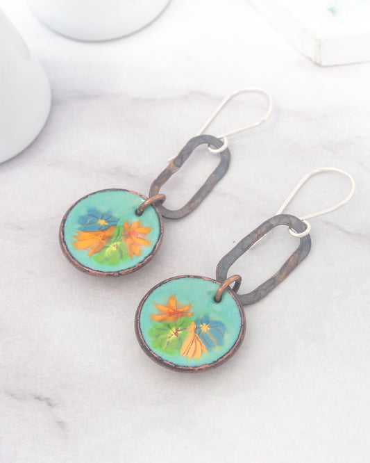 A pair of earrings with a floral design on the circular pendants, resting on a marble surface.