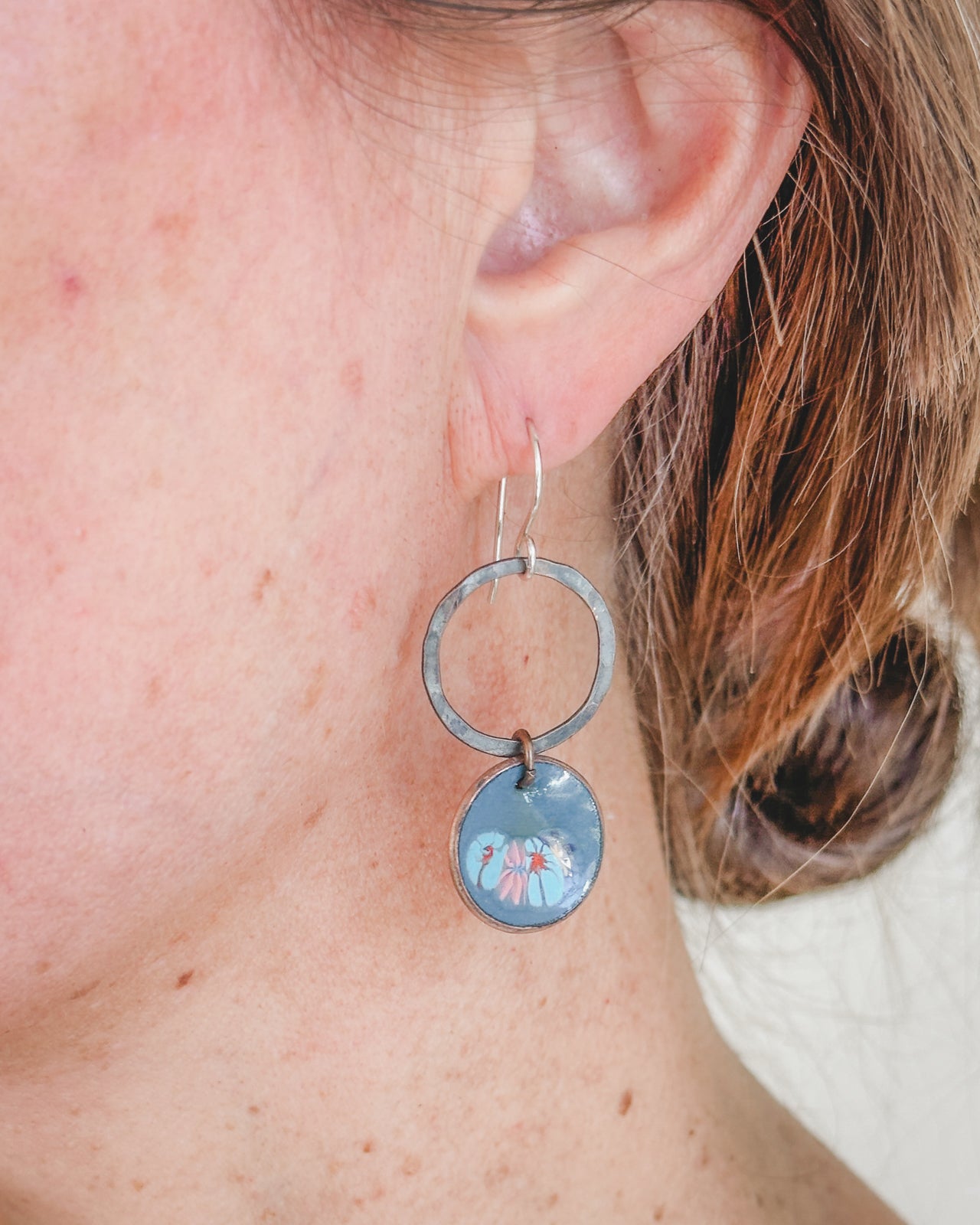 A close-up view of a person's ear wearing a pair of silver earrings with a blue circular charm featuring a floral design.