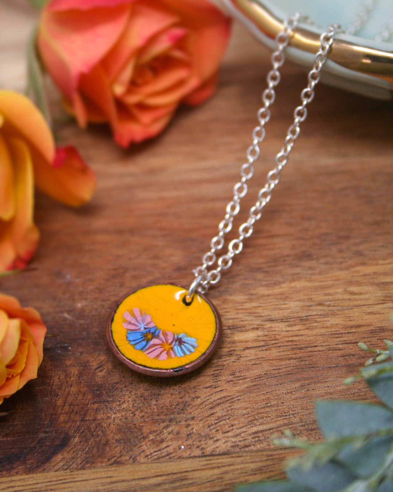 A necklace with a circular pendant featuring a floral design is displayed on a wooden surface, surrounded by orange and pink roses.