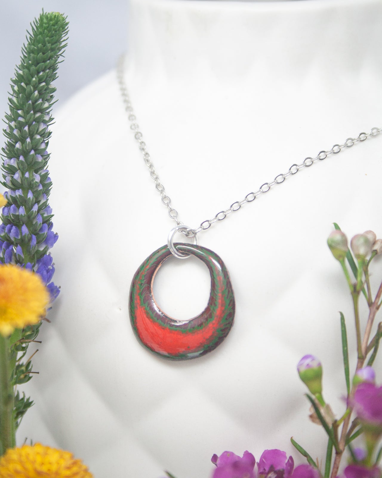 A necklace with a colorful pendant is displayed in a white vase, surrounded by various flowers.