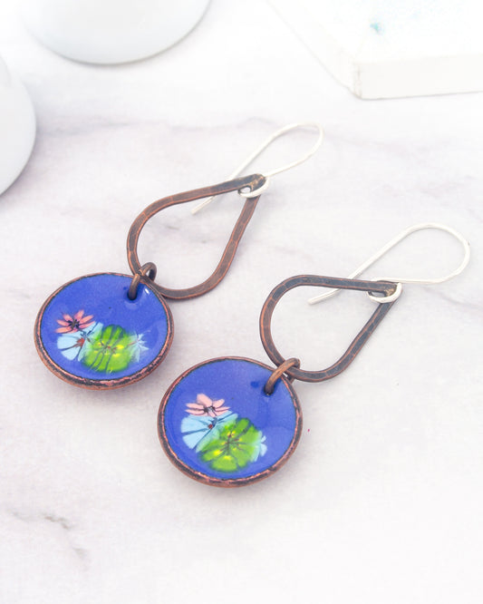 A pair of earrings with blue and green floral designs on circular blue discs, hanging from metal hooks.