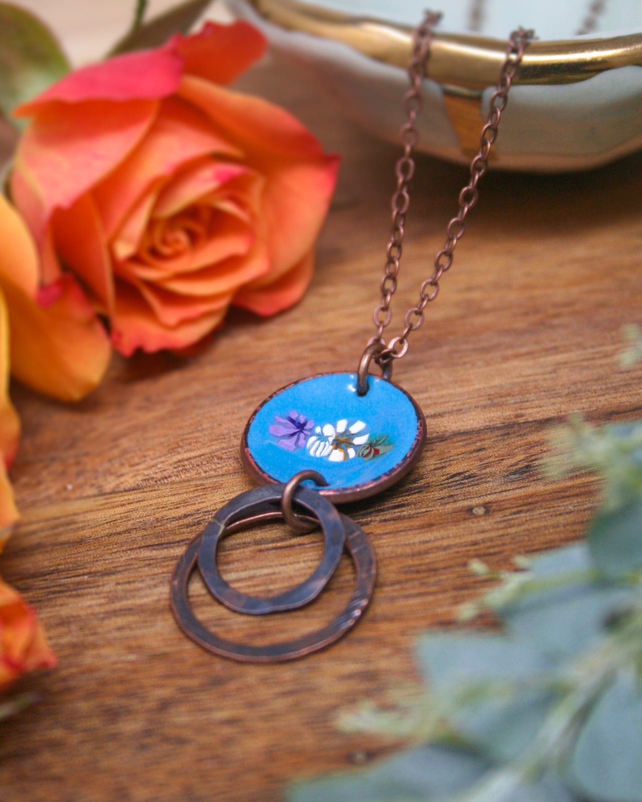 A blue pendant necklace with a circular design and two circular rings is displayed on a wooden surface, accompanied by a vibrant orange rose and some greenery.