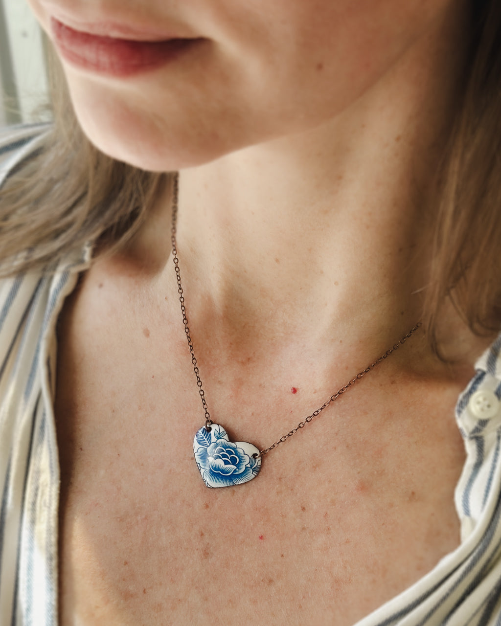 A close-up of a woman's neck and chest area, showing a delicate heart-shaped pendant necklace with a blue floral design.