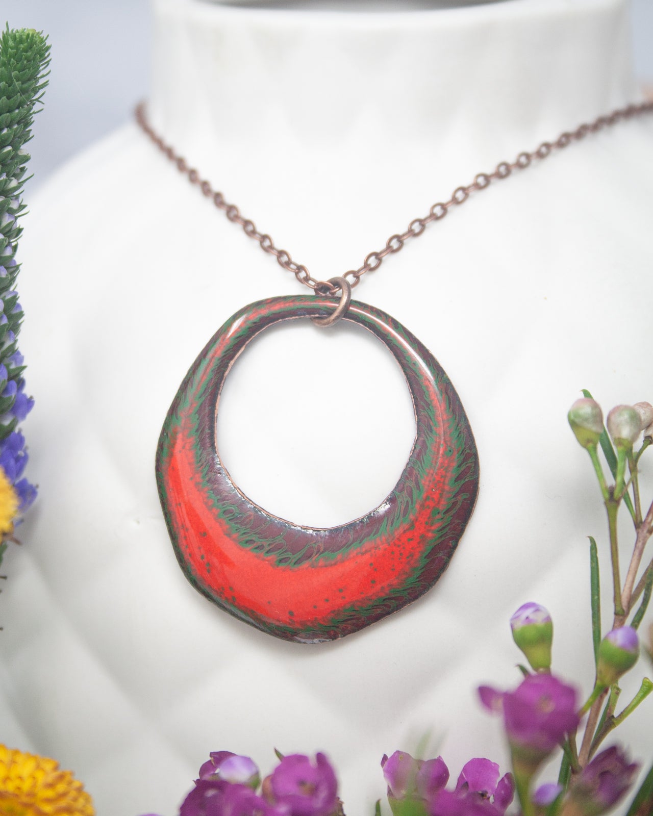 A necklace with a large, colorful pendant is displayed against a white background, surrounded by flowers.