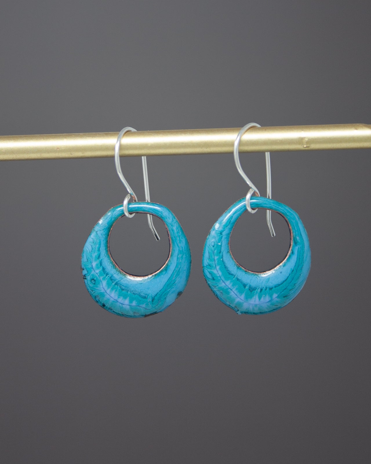 A pair of turquoise-colored hoop earrings with a metallic finish, hanging from a gold-colored metal bar.