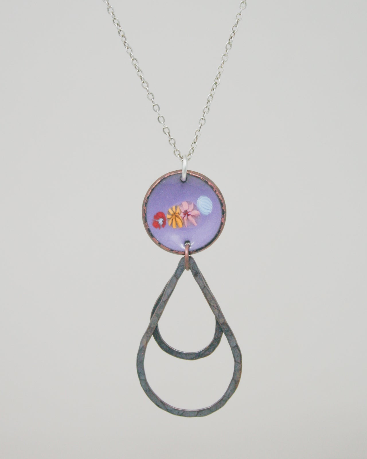 A necklace with a purple pendant featuring a circular design with a floral pattern and a teardrop-shaped metal element hanging from it.