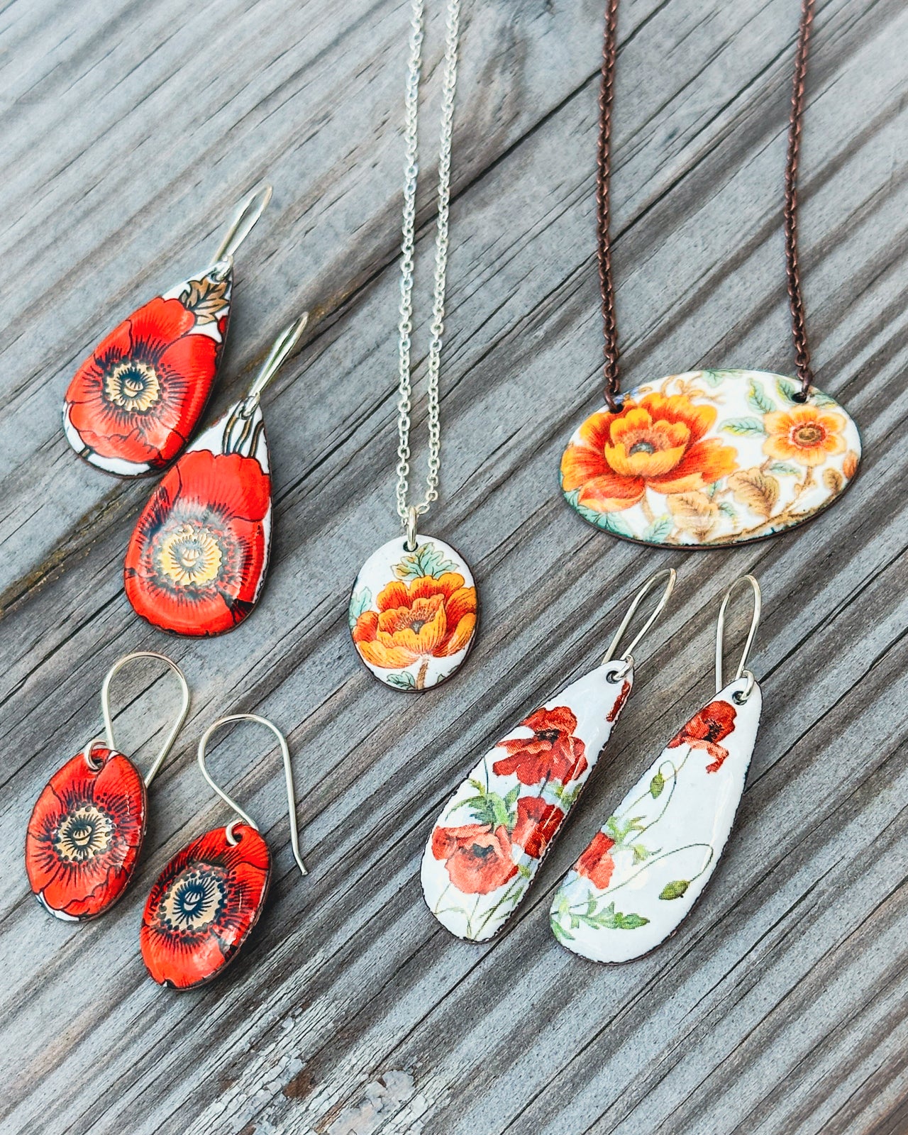 a collection of colorful earrings and necklaces featuring floral designs, including red poppies, on a wooden surface.