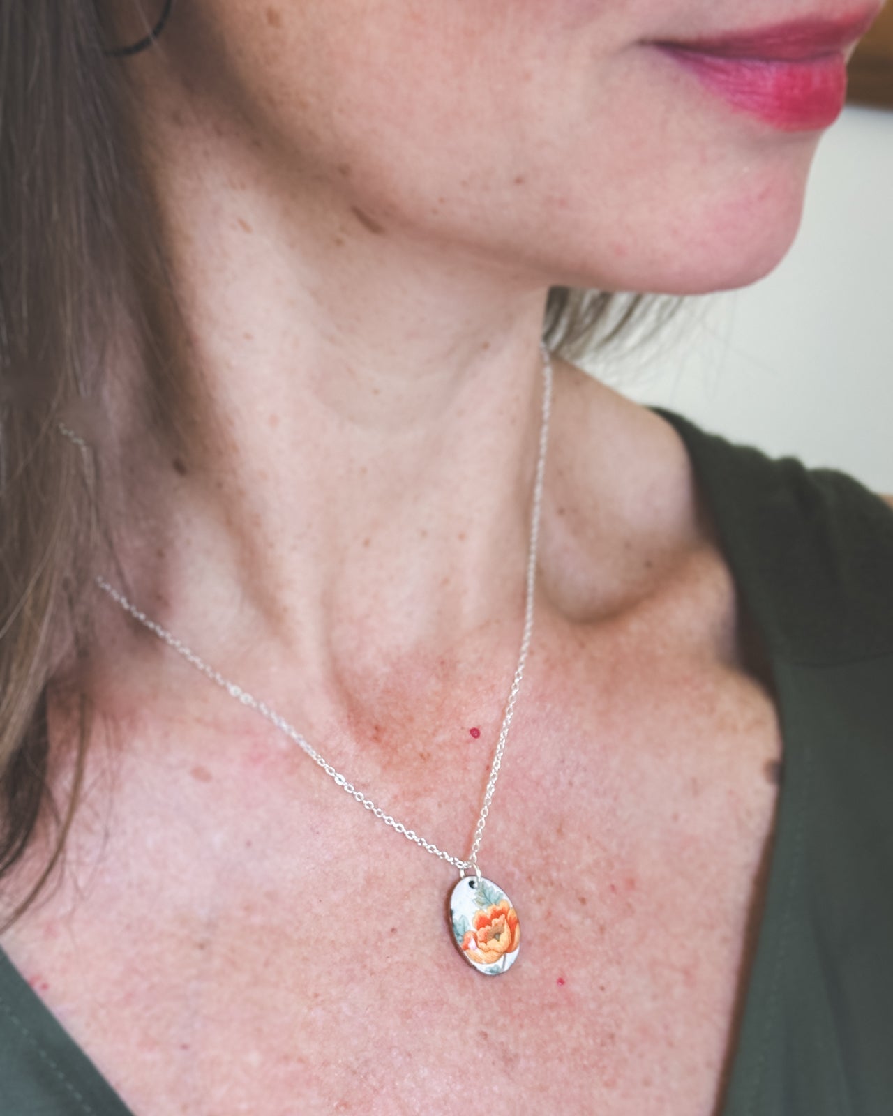 A close-up of a woman's neck and chest area, with a silver necklace featuring a pendant with an orange flower design.