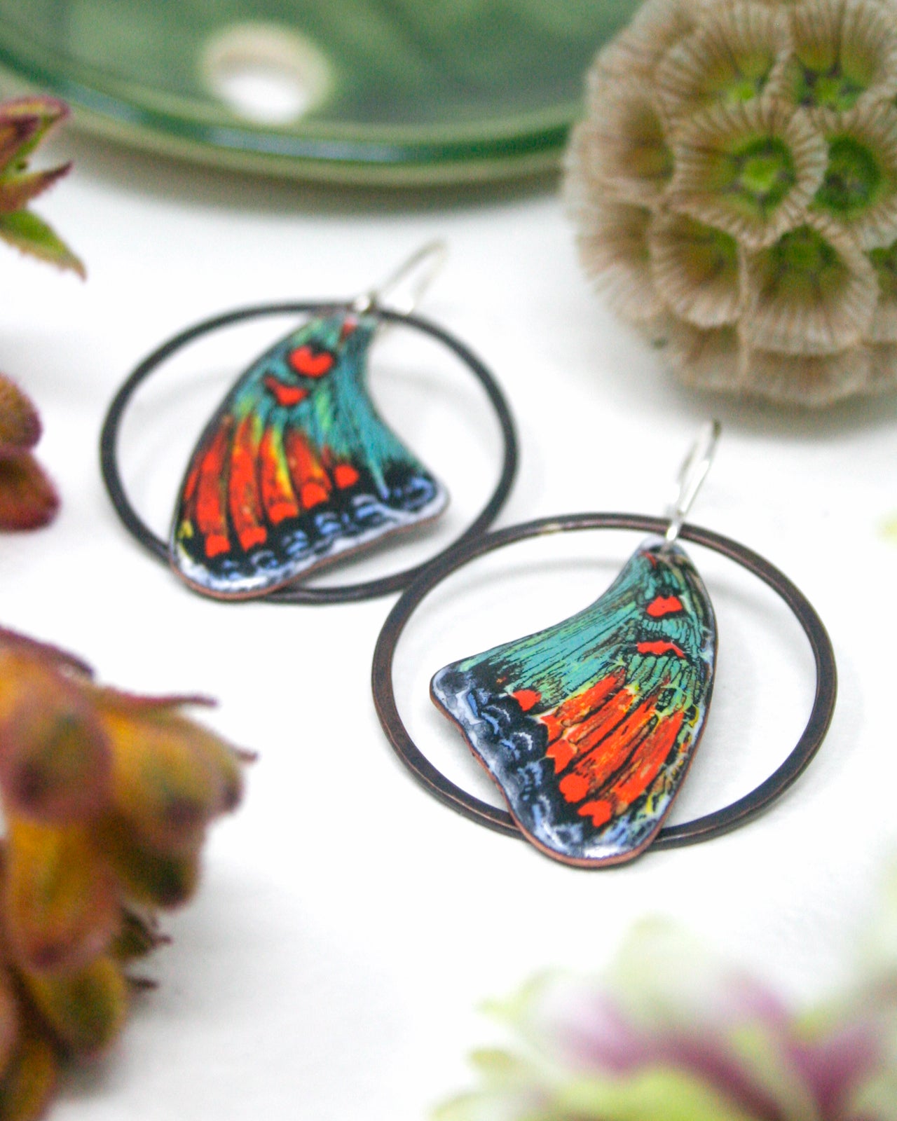 a pair of colorful butterfly-shaped earrings with intricate designs, placed on a white surface alongside some flowers.