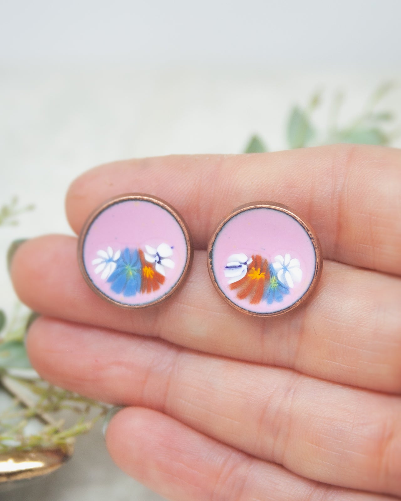 A hand holding two circular earrings with colorful floral designs on them.