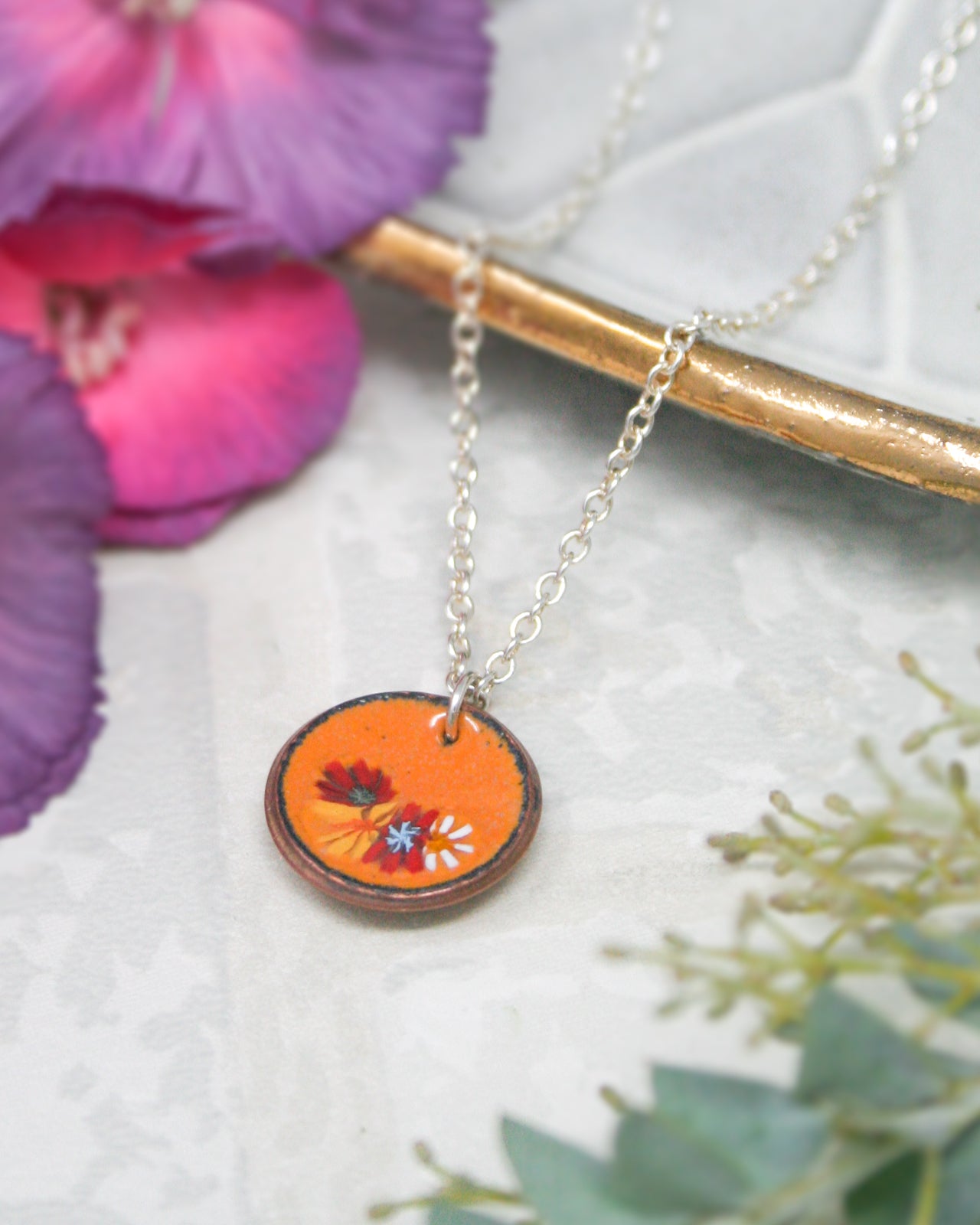 A necklace with a circular pendant featuring a floral design is displayed on a white surface, surrounded by purple and green flowers.