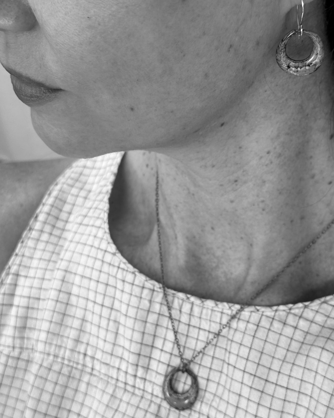 A close-up black and white image of a woman's neck and chest area, showing a necklace with a pendant and a pair of earrings.