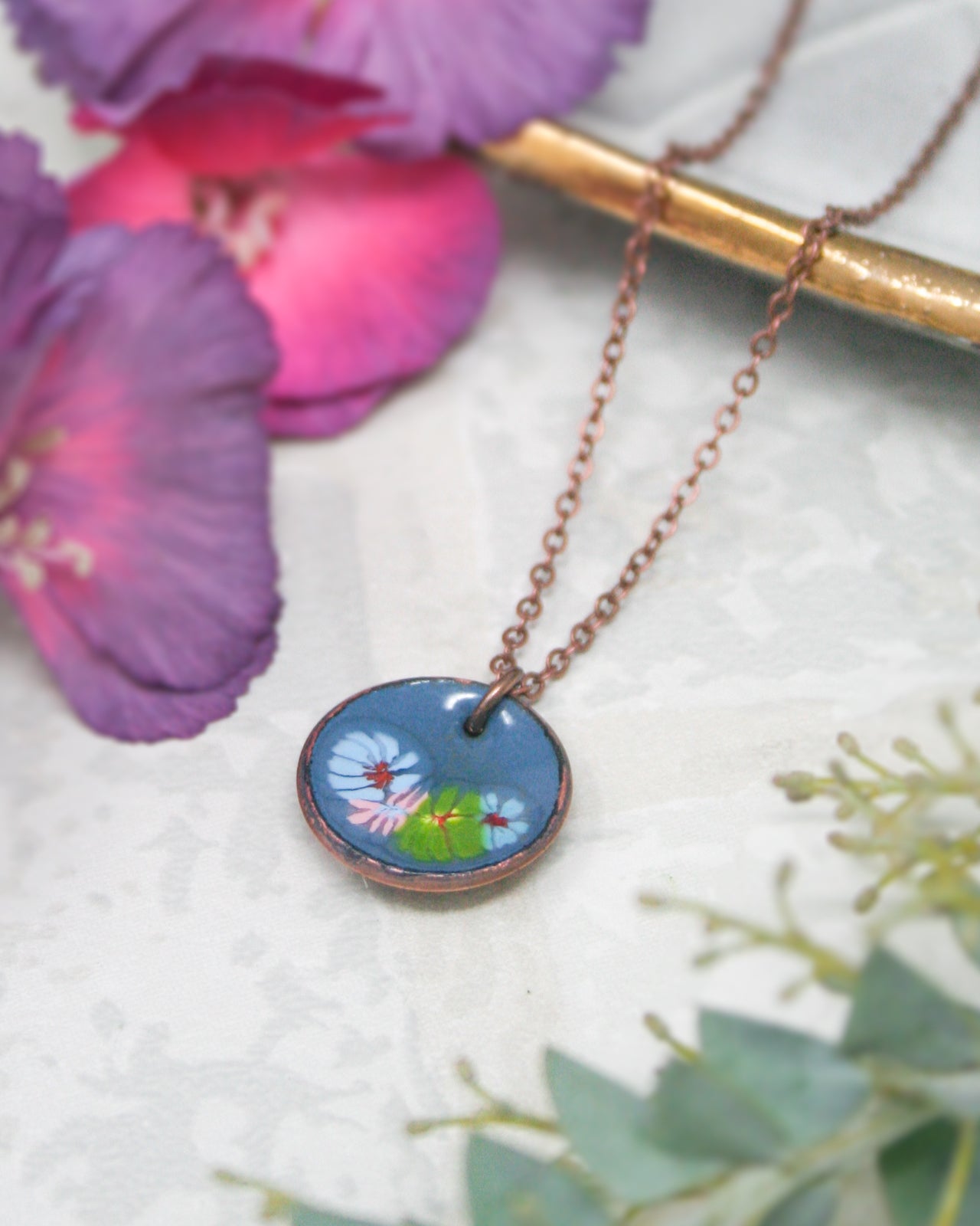 A necklace with a blue pendant featuring a floral design is displayed, surrounded by purple and pink flowers and a gold-colored metal object.