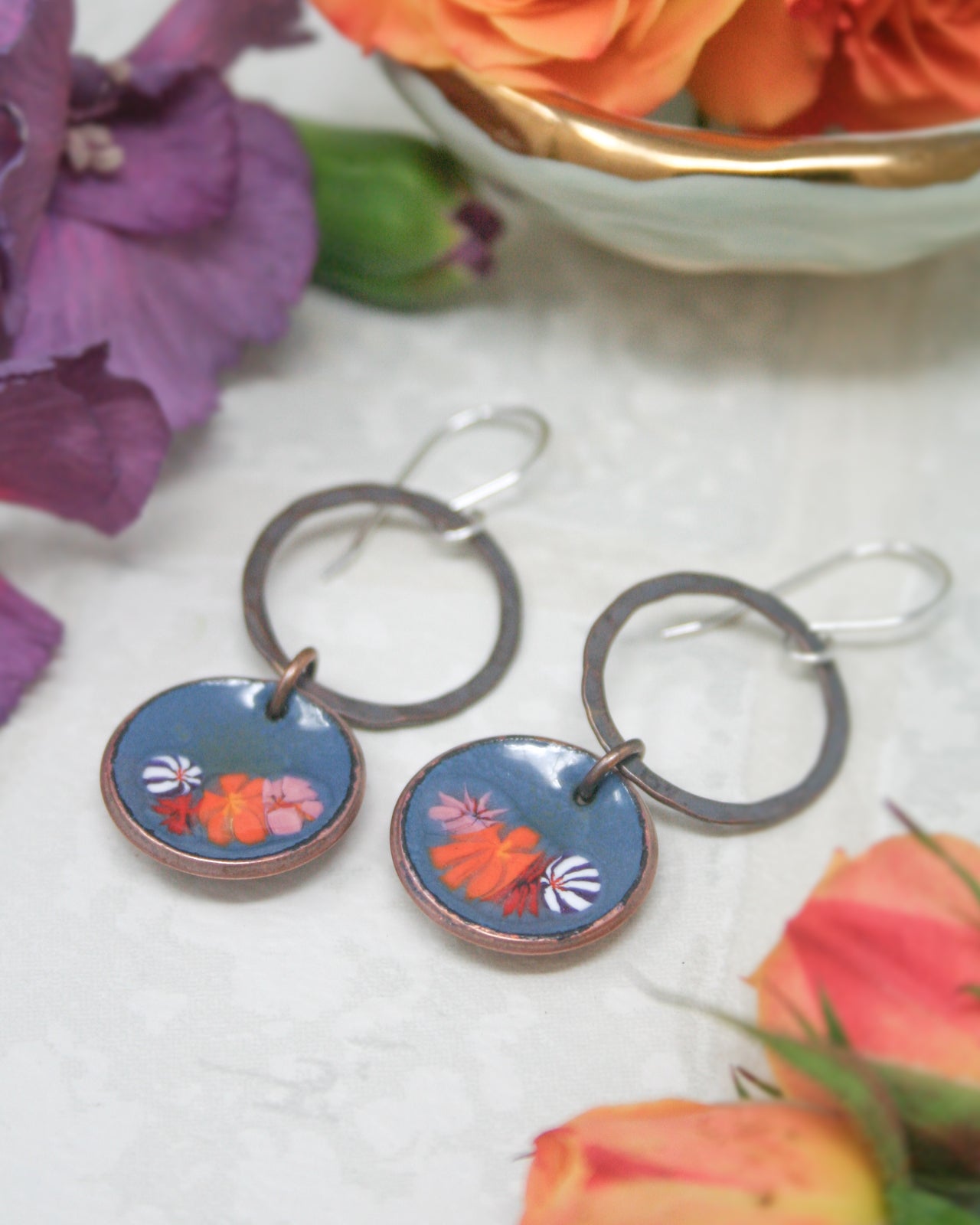 a pair of blue and orange earrings with floral designs, placed on a white surface alongside a bowl of orange flowers and purple flowers.