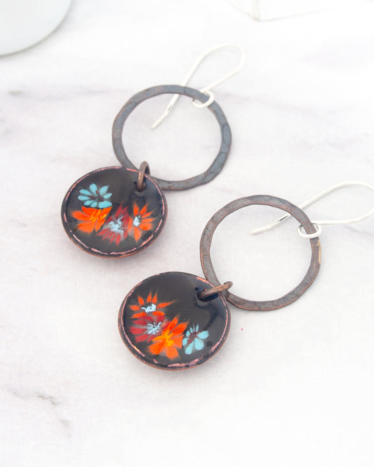 A pair of earrings with a floral design on the circular pendants, resting on a white marble surface.