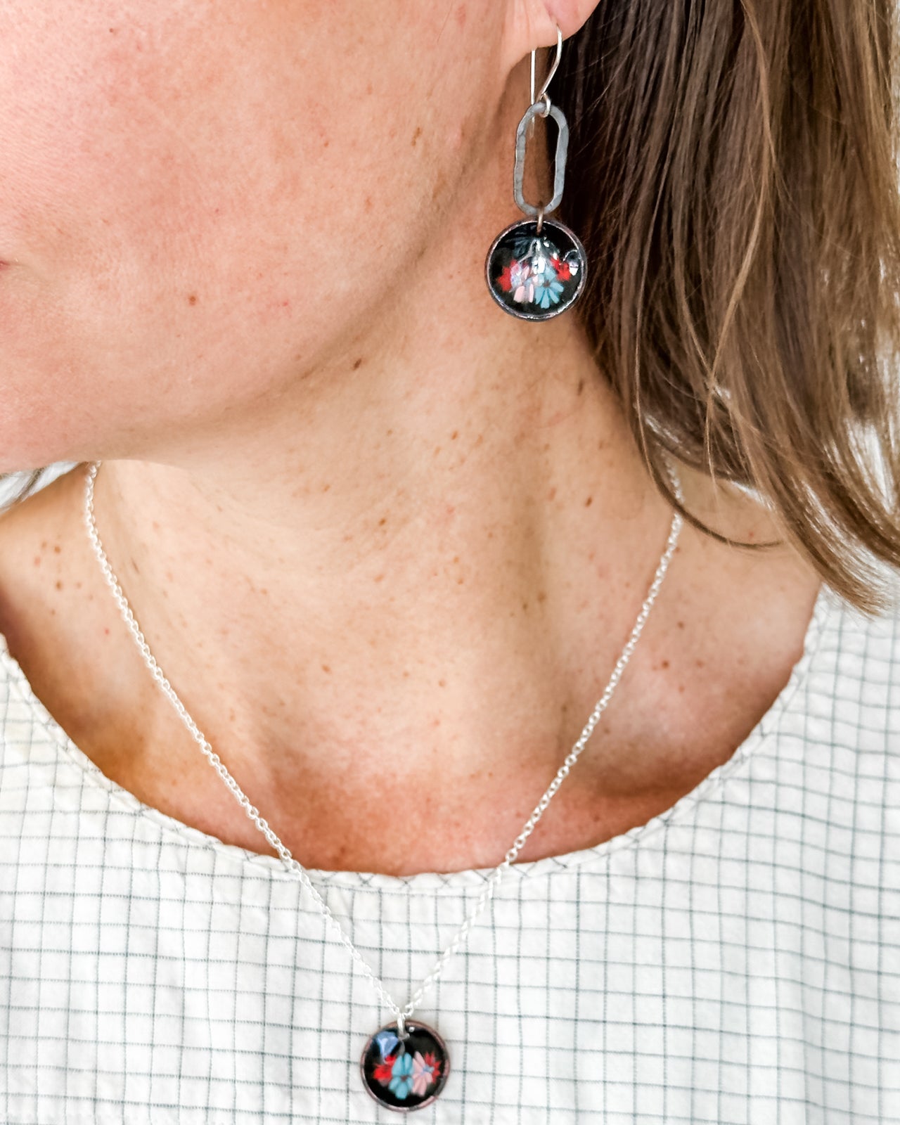 A woman wearing a necklace with a pendant and earrings, and a white shirt with a plaid pattern.
