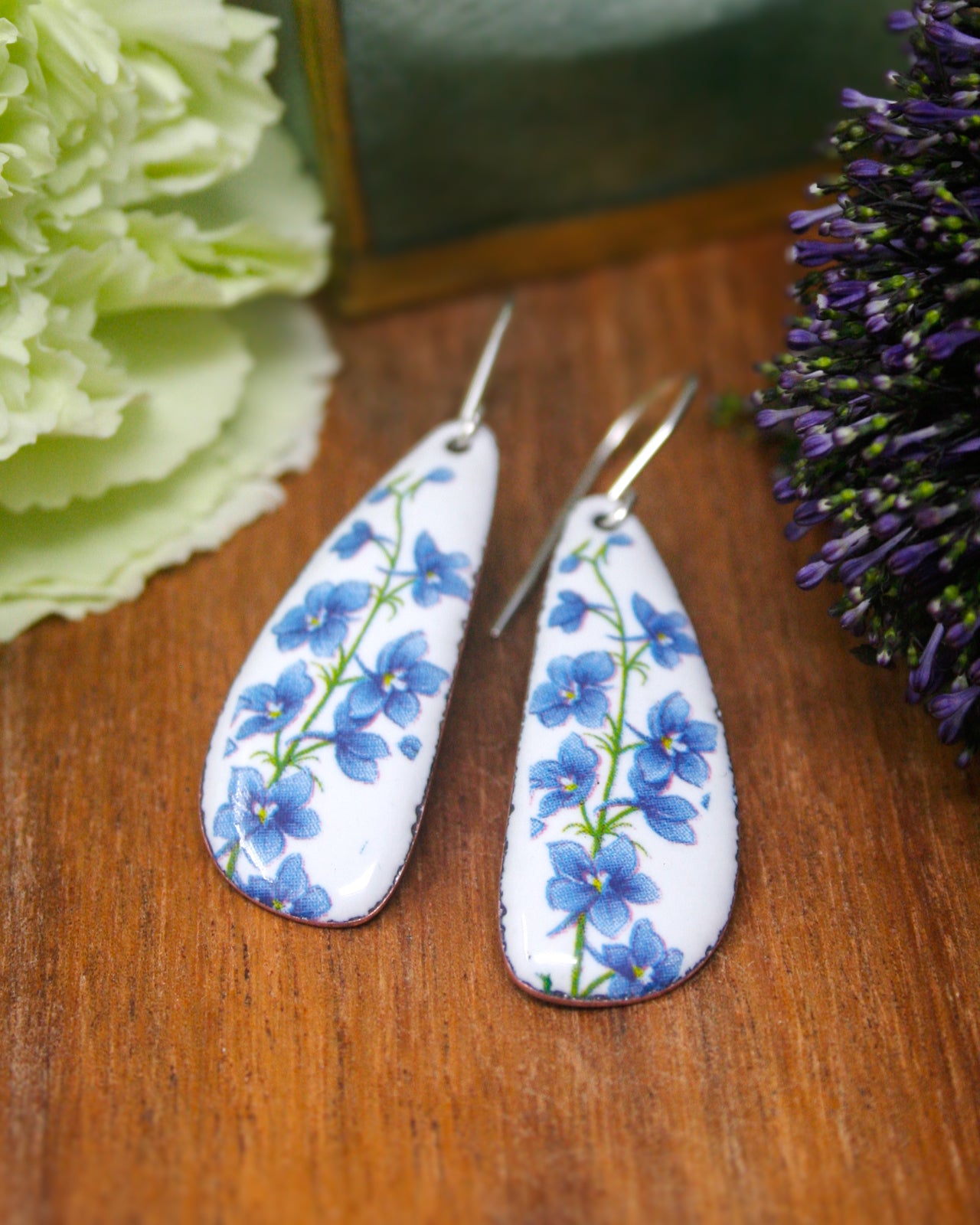 A pair of white earrings with blue flower designs rests on a wooden surface, accompanied by a green flower and purple flowers.