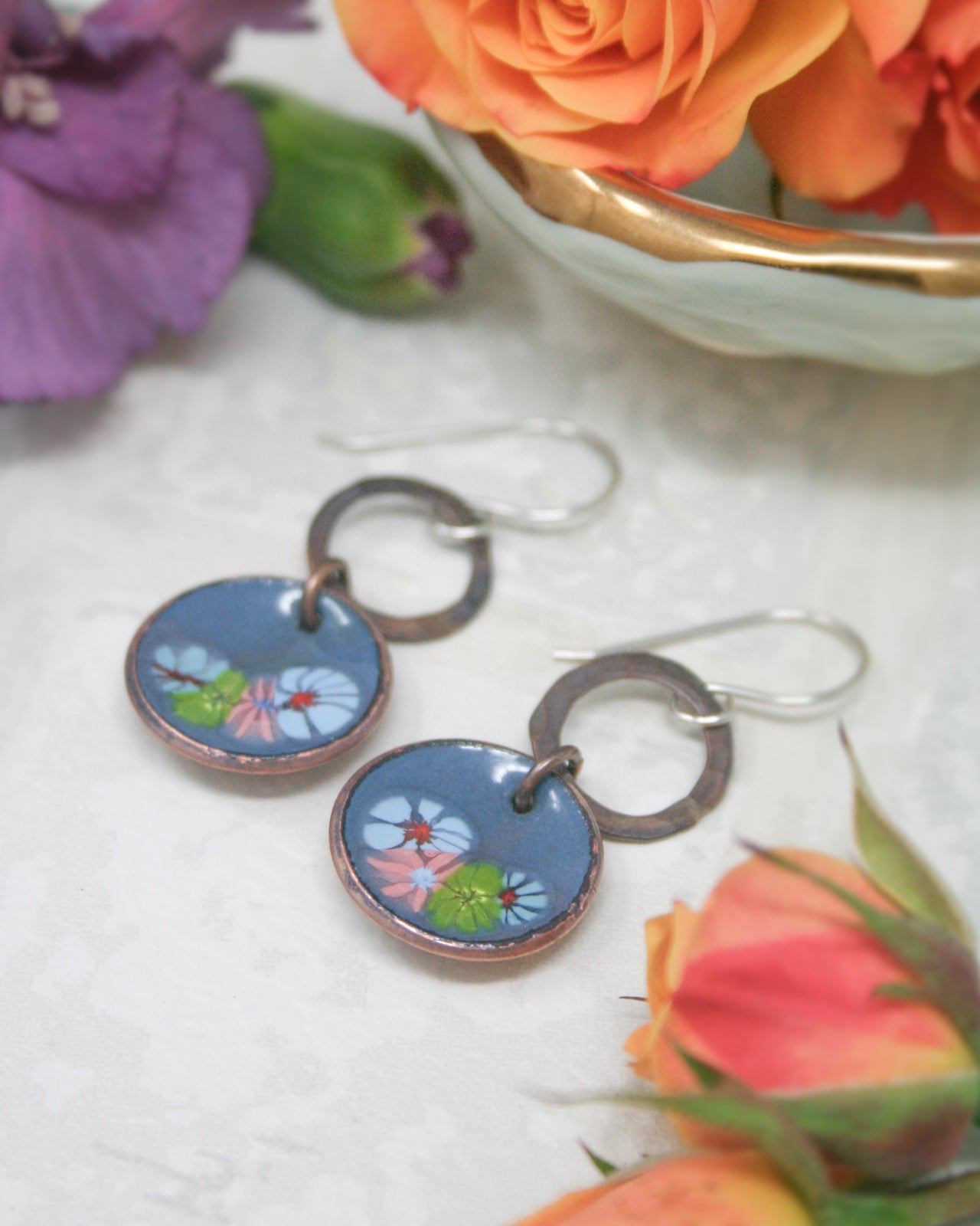 a pair of blue and copper earrings with floral designs, placed on a white surface alongside a bowl of orange and purple flowers.