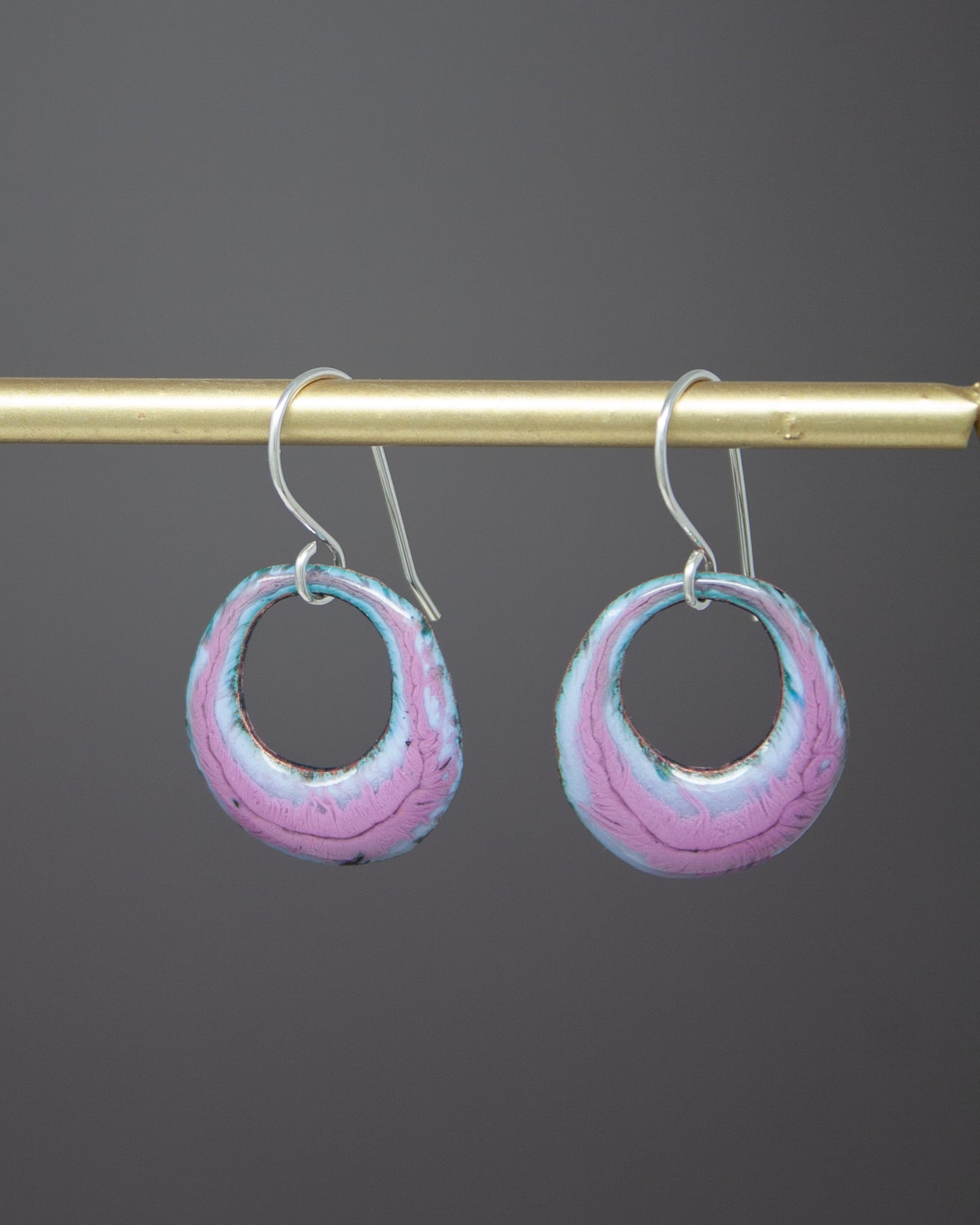 A pair of earrings with a circular design, featuring a combination of pink and blue colors. The earrings are hanging from a gold-colored metal bar or hook.
