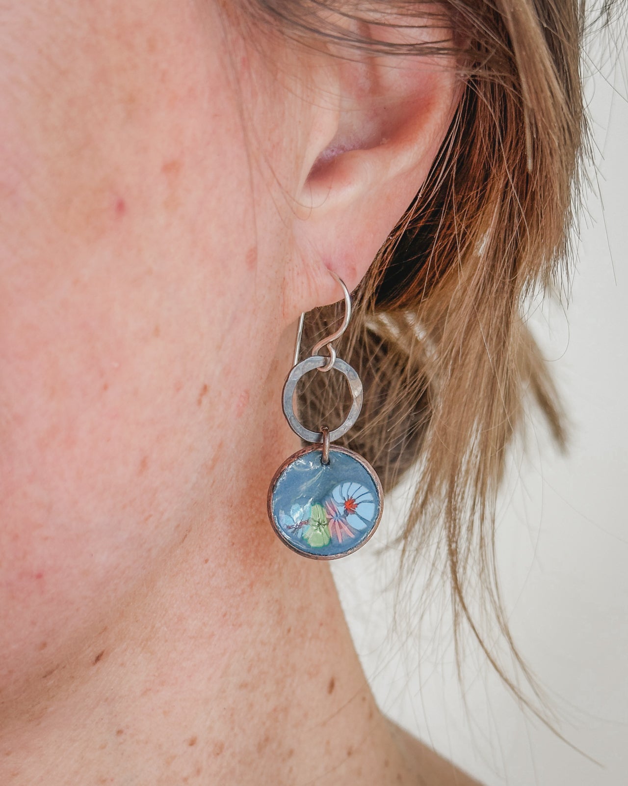 A close-up view of a person's ear wearing a pair of silver earrings with a blue floral design.