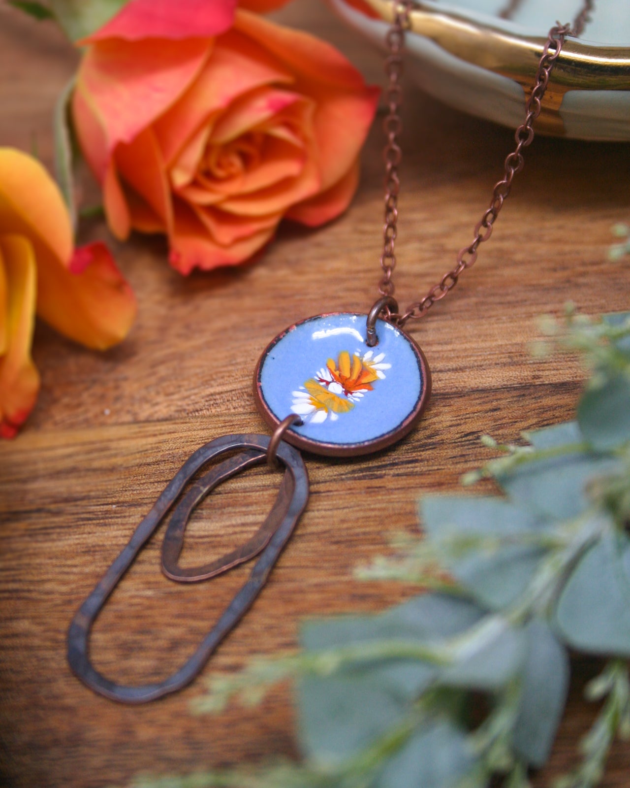 a necklace with a blue pendant adorned with a floral design, resting on a wooden surface. The necklace is accompanied by a pair of orange roses and some greenery, creating a visually appealing and harmonious composition.