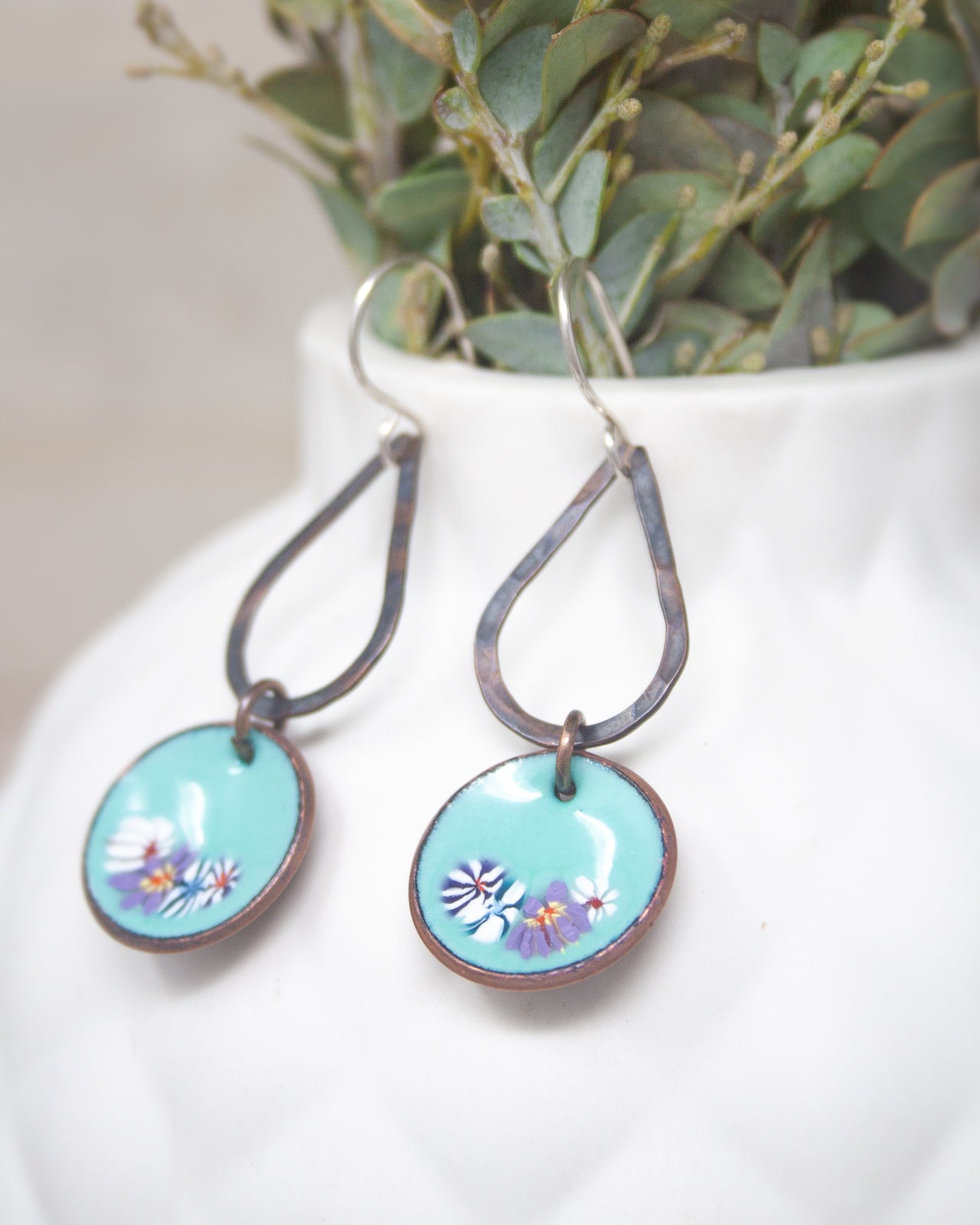 A pair of turquoise earrings with floral designs, hanging from hooks in a white vase.