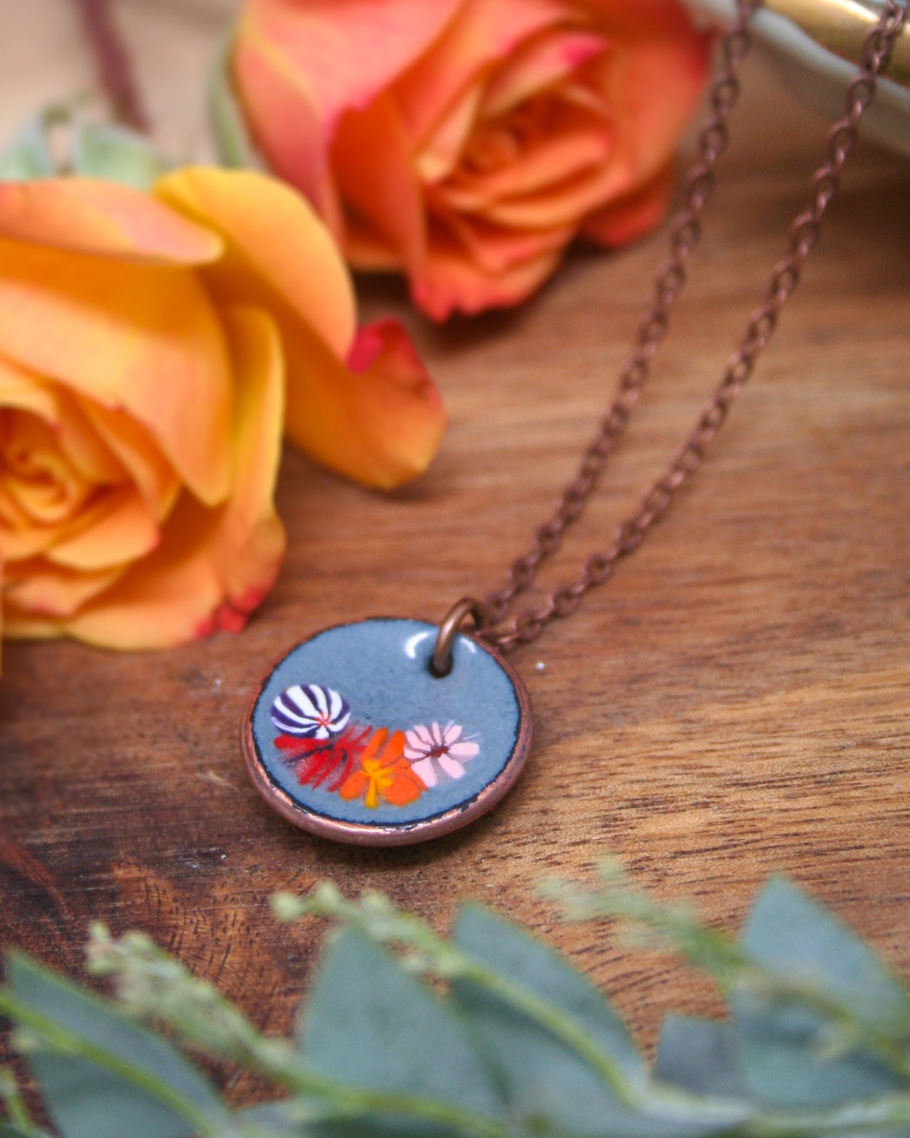 A pendant necklace with a circular design featuring colorful flowers rests on a wooden surface, surrounded by vibrant orange and yellow roses.
