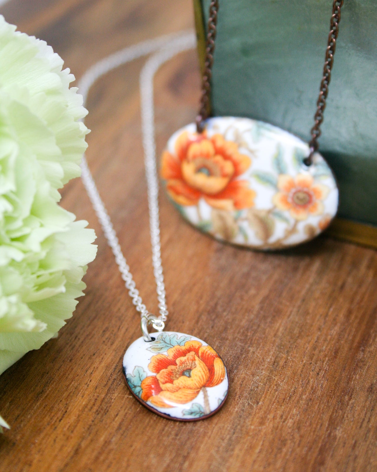 a necklace with two pendants, one of which is a floral design in orange and white colors, and the other is a white oval pendant with a floral design. The pendants are placed on a wooden surface, with a green object in the background.