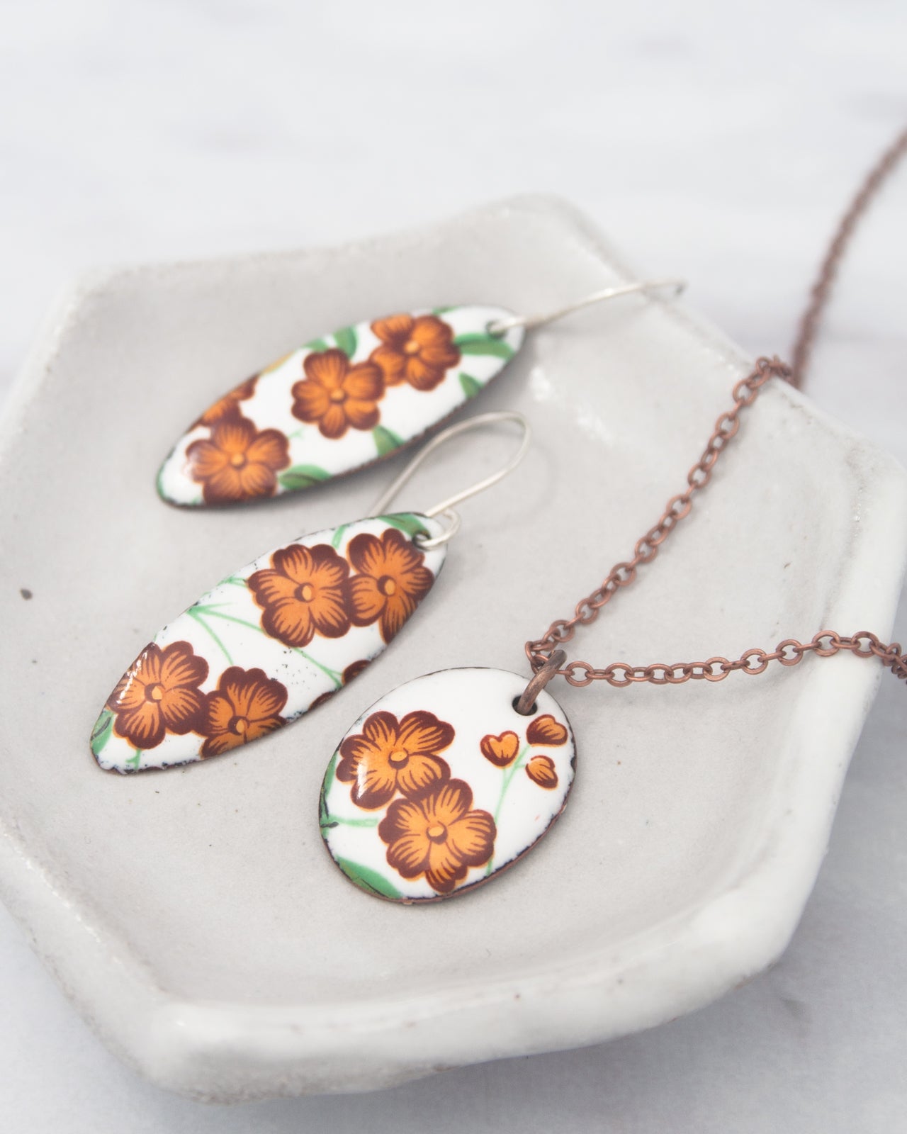 a pair of earrings and a necklace with floral designs, including orange flowers, displayed on a white plate.