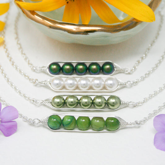 a collection of necklaces with various colored beads, including green, white, and purple beads, arranged in a row. The necklaces are displayed on a white surface, with a bowl containing yellow flowers in the background.
