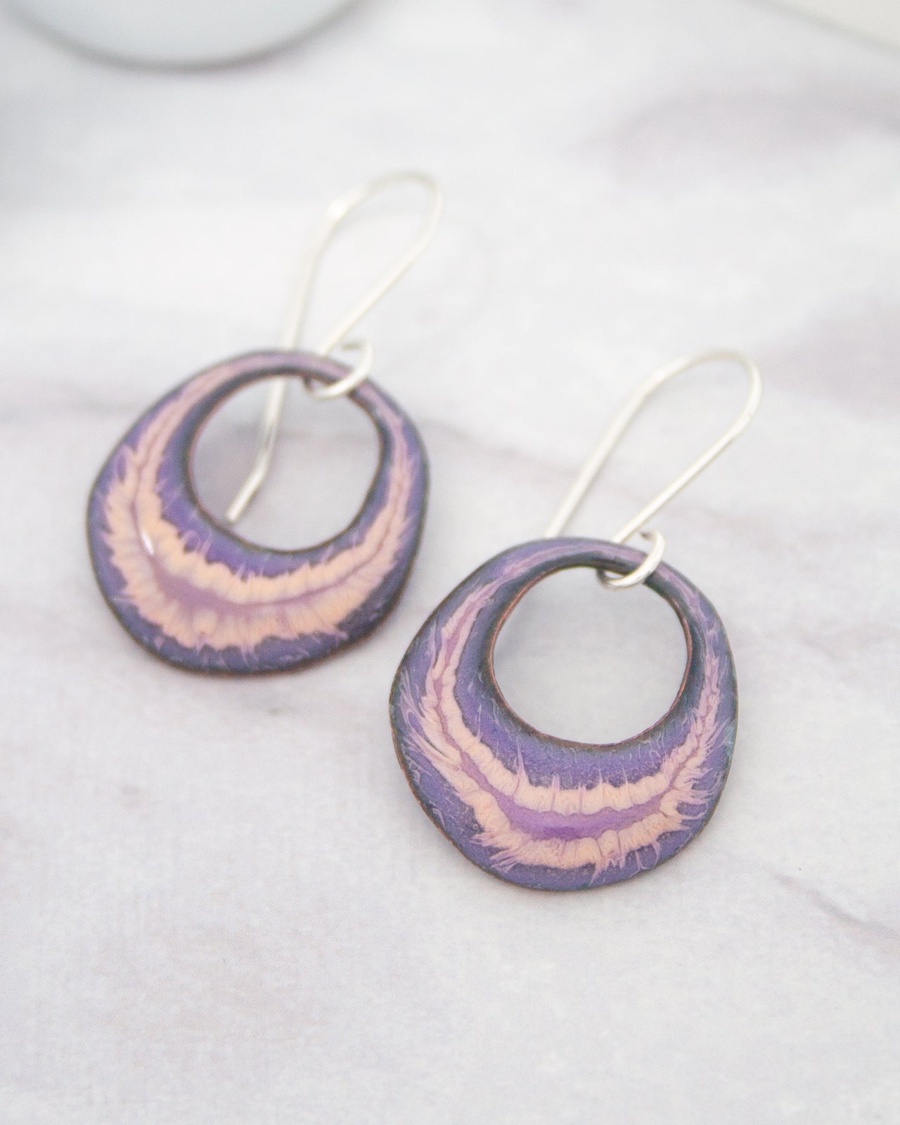 A pair of earrings with a purple and pink color scheme, featuring circular designs with a metallic finish.