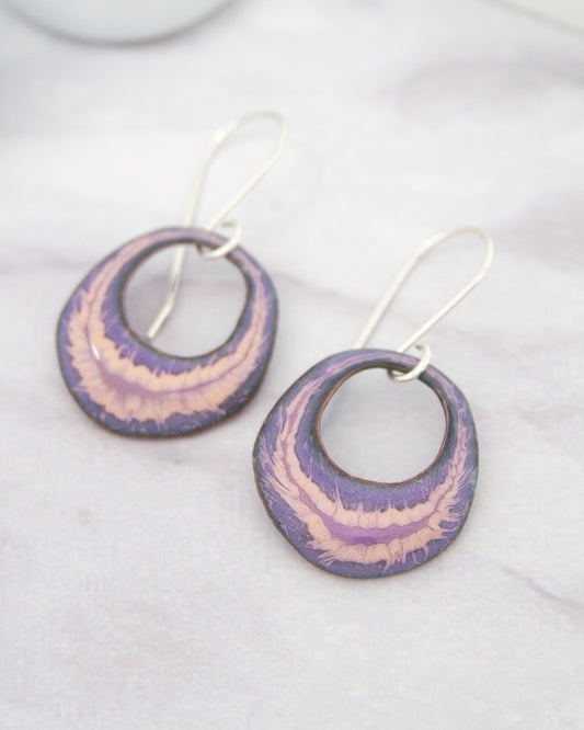 A pair of earrings with a purple and pink color scheme, featuring circular designs with a metallic finish.