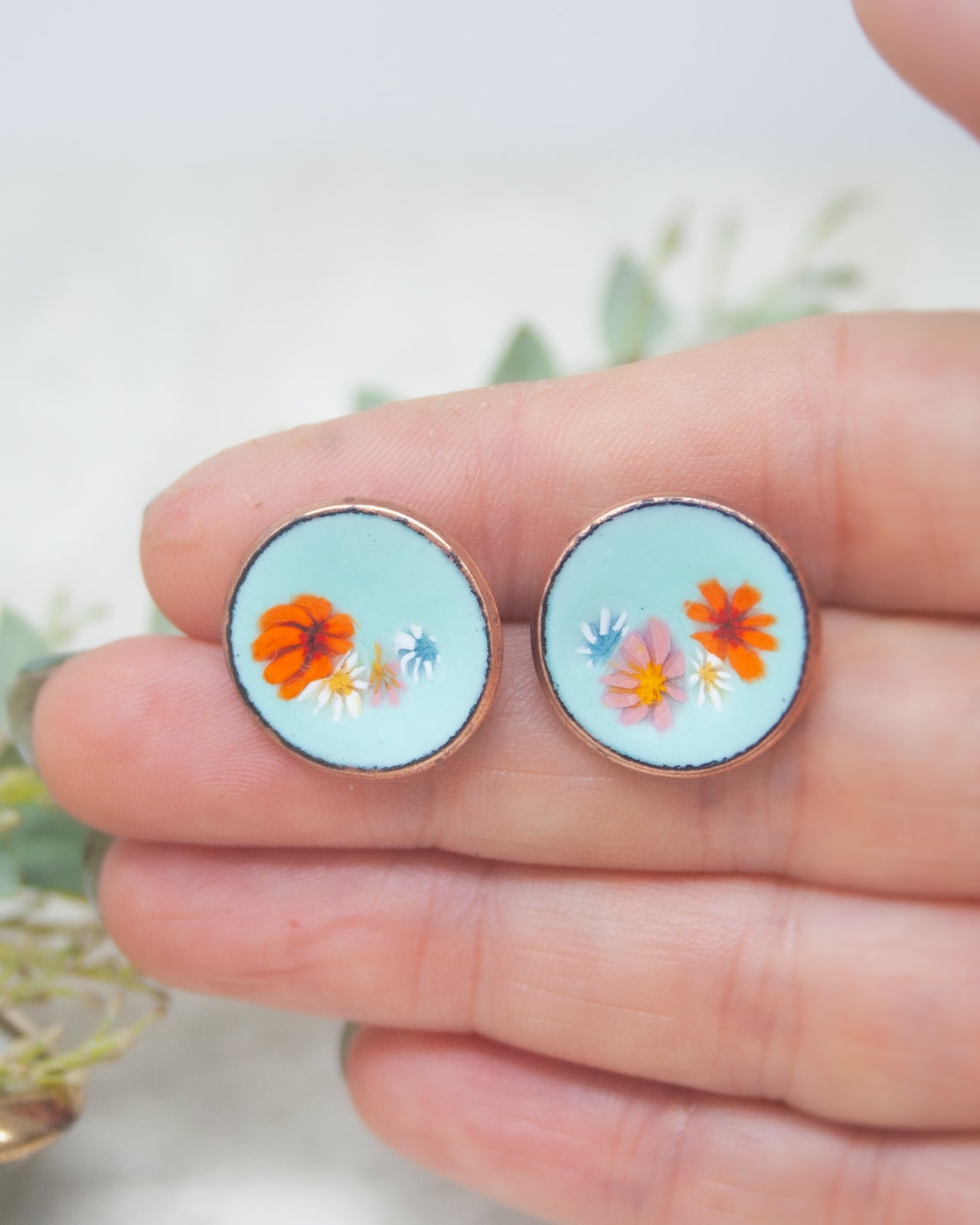 A hand holding a pair of blue earrings with colorful flower designs.