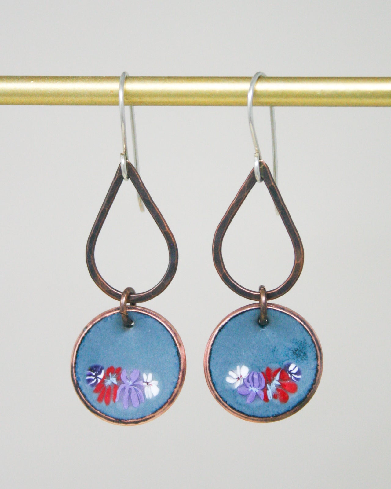 A pair of earrings with blue and purple floral designs on circular blue discs, hanging from a gold-colored metal hook.
