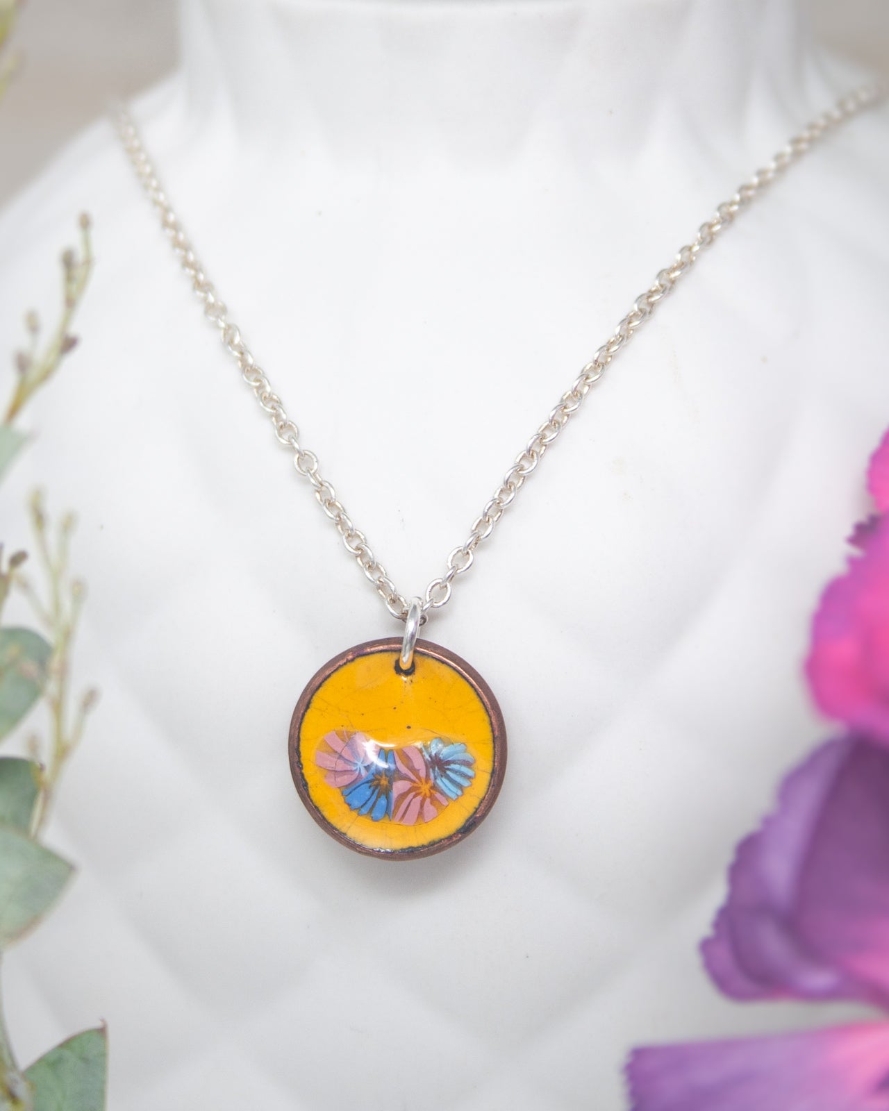 A necklace with a circular pendant featuring a floral design in yellow and purple colors.