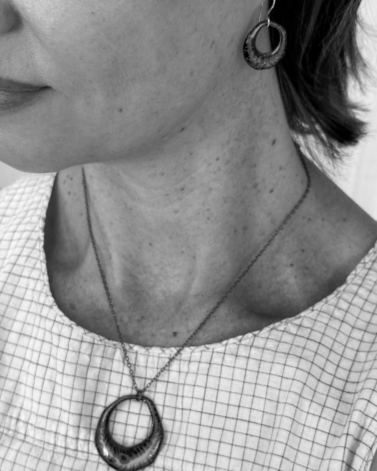 A close-up black and white image of a woman wearing a necklace with a pendant and earrings.
