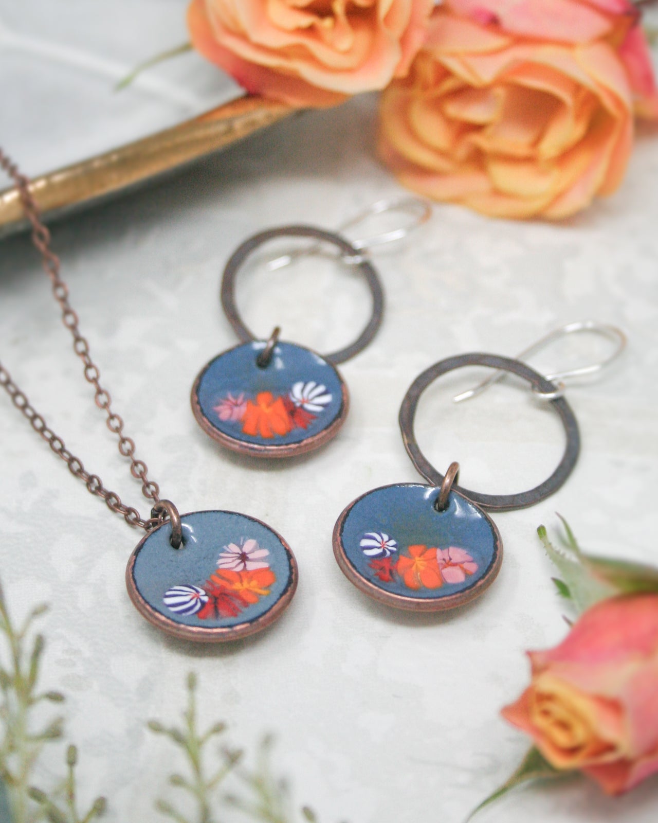 a necklace with a pendant and earrings with a floral design, all placed on a white surface with orange and pink roses in the background.