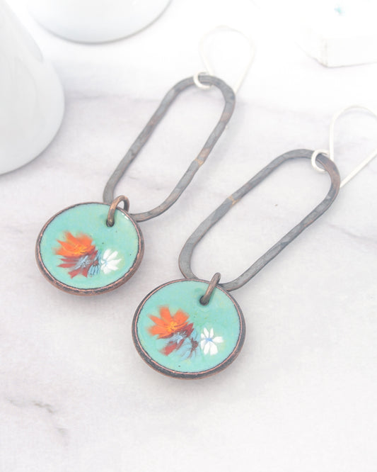 A pair of earrings with a floral design on the circular pendants, resting on a white surface.