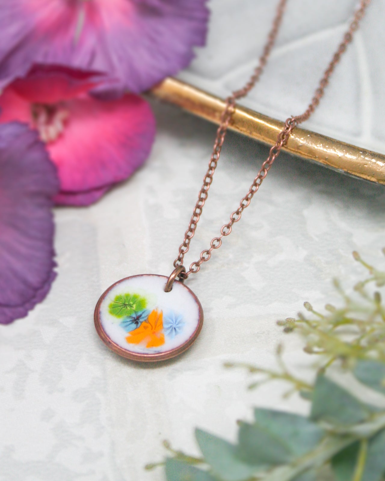 A necklace with a circular pendant featuring colorful flowers is displayed on a white surface, with a purple flower and a gold-colored object in the background.