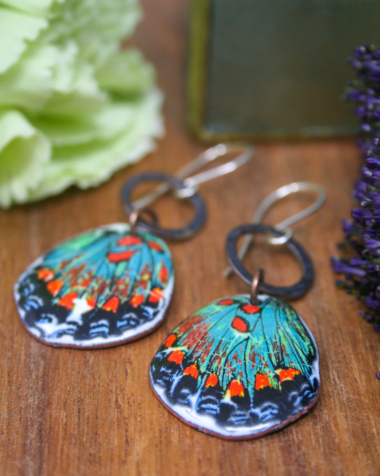 a pair of colorful earrings with butterfly designs, placed on a wooden surface alongside some purple flowers.