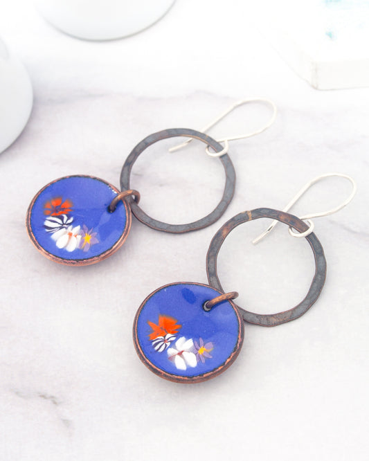 A pair of earrings with blue and orange floral designs on circular blue backgrounds, hanging from silver hooks.