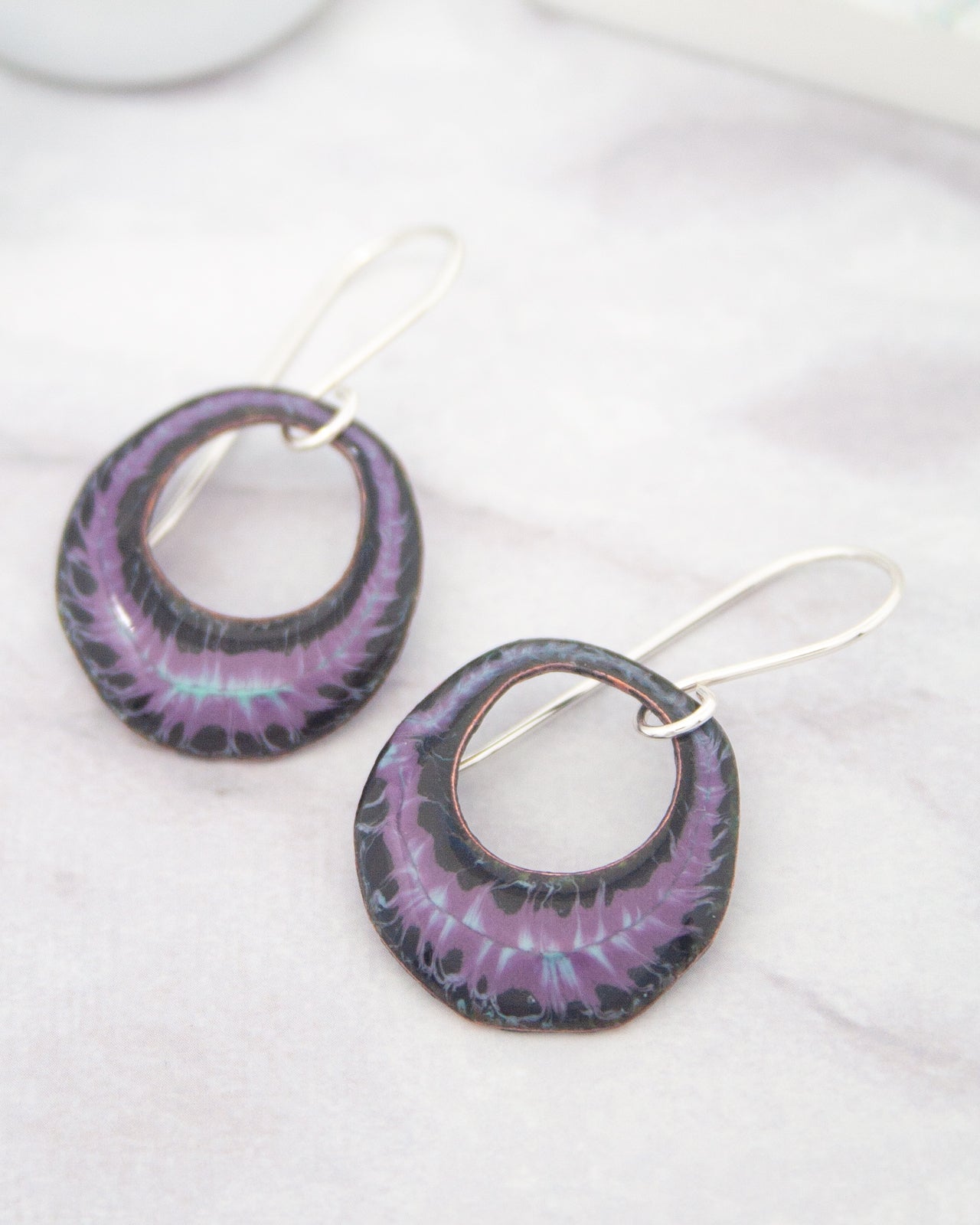 A pair of earrings with purple and black metallic designs, featuring circular cutouts and a metallic chain.