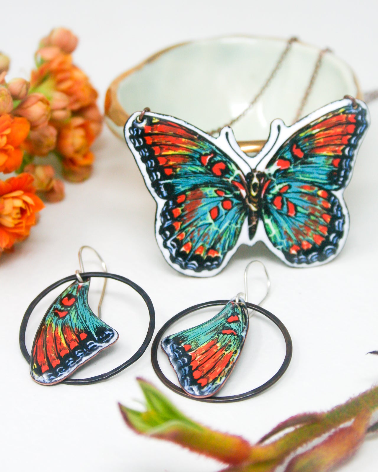 a pair of colorful butterfly-themed earrings and a butterfly-shaped necklace, along with some orange flowers and a white bowl.